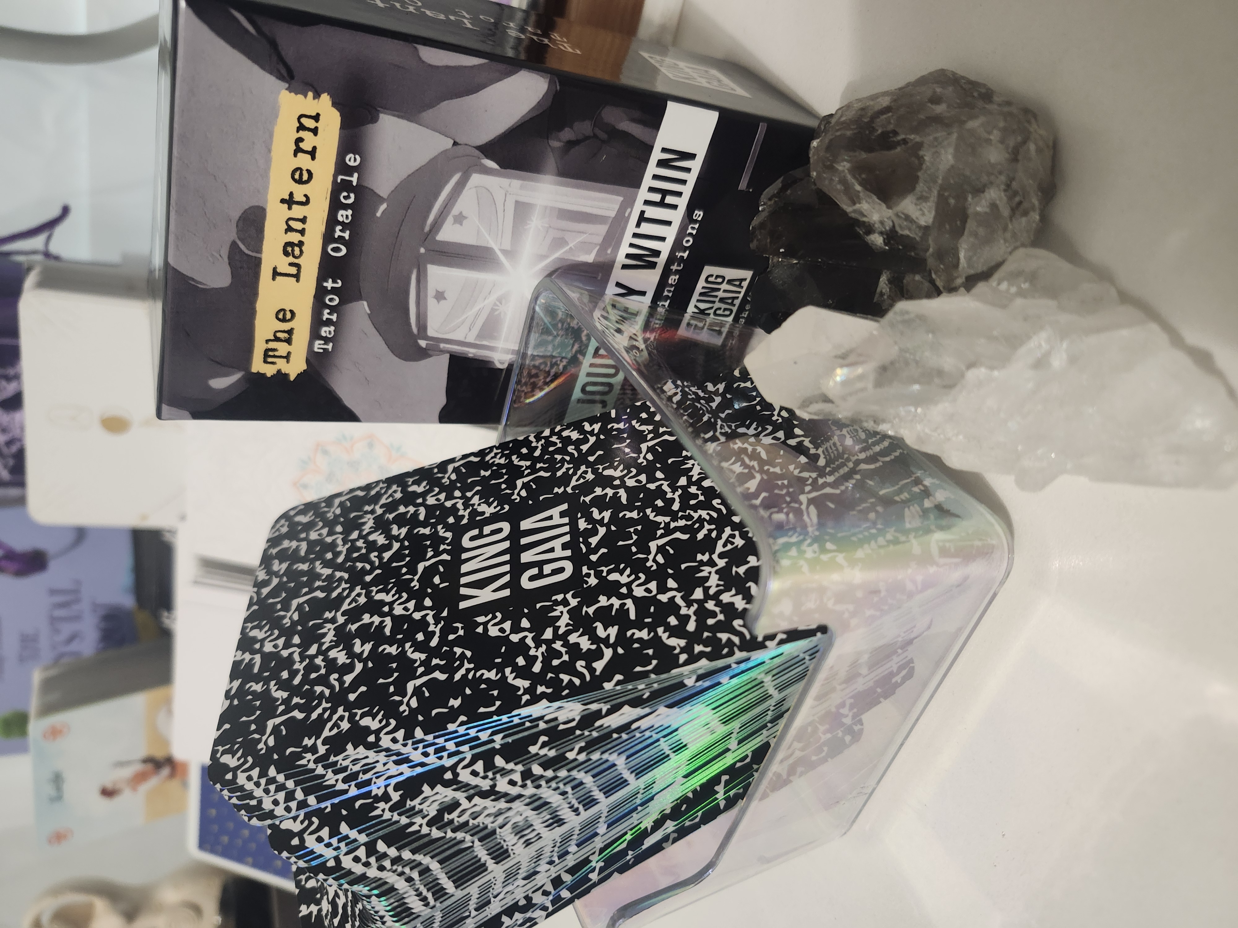 The Lantern Tarot Oracle deck by King Gaia fanned open to reveal holographic edges and the Journey Within Illuminations box surrounded by healing crystals. Designed for introspection and transformation.